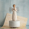 Angel figurine in white dress with hands folded in prayer - she has black wire wings and is standing on round white plaque - background is light blue with a tan colored music sheet below her