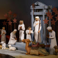 A carved nativity scene set out on a wood table laid with a linen cloth and lit candles in the background.