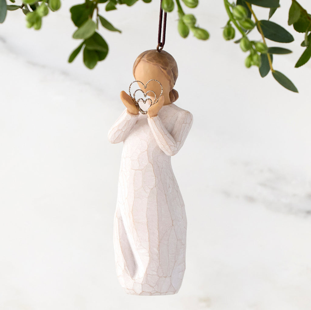 Figure in cream dress, holding thin wire concentric heart in hands. Wire heart is dusted with gold glitter. Hook and loop affixed to figure's head