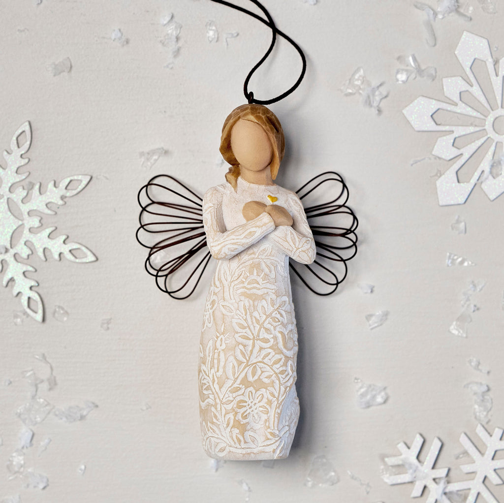 Willow Tree Remembrance Ornament, 4.5”h hand-painted resin angel figure in cream dress carved with symbols of nature, wire wings, crossed hands under small gold-leaf heart, brown elastic cord, laying amongst snowflakes