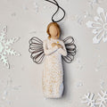 Willow Tree Remembrance Ornament, 4.5”h hand-painted resin angel figure in cream dress carved with symbols of nature, wire wings, crossed hands under small gold-leaf heart, brown elastic cord, laying amongst snowflakes