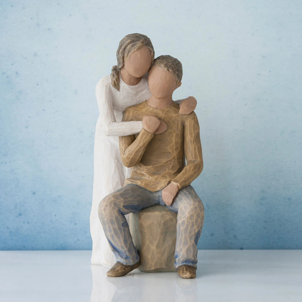 Man figurine sitting on rock wearing brown shirt and blue jeans with woman figurine in white dress with arms wrapped around him - blue background
