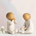 Willow Tree Two Together, two hand-painted seated resin figures of babies, tallest 2", one with hands at mouth, one with hands on legs, both in cream onsies, in front of pastel painting