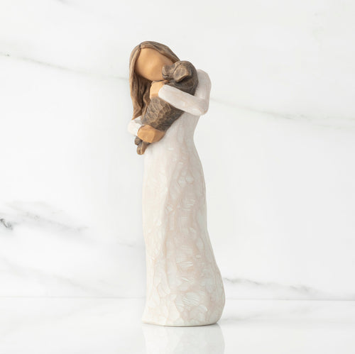 Left view, standing female figure in cream dress, holding dark color puppy in her arms