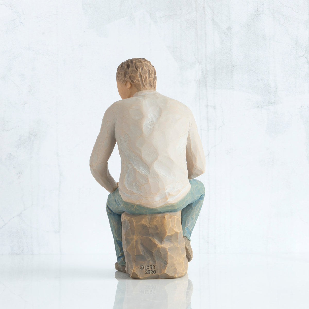 Back view, adult male figure in light gray shirt and blue jeans, seated on gray rock