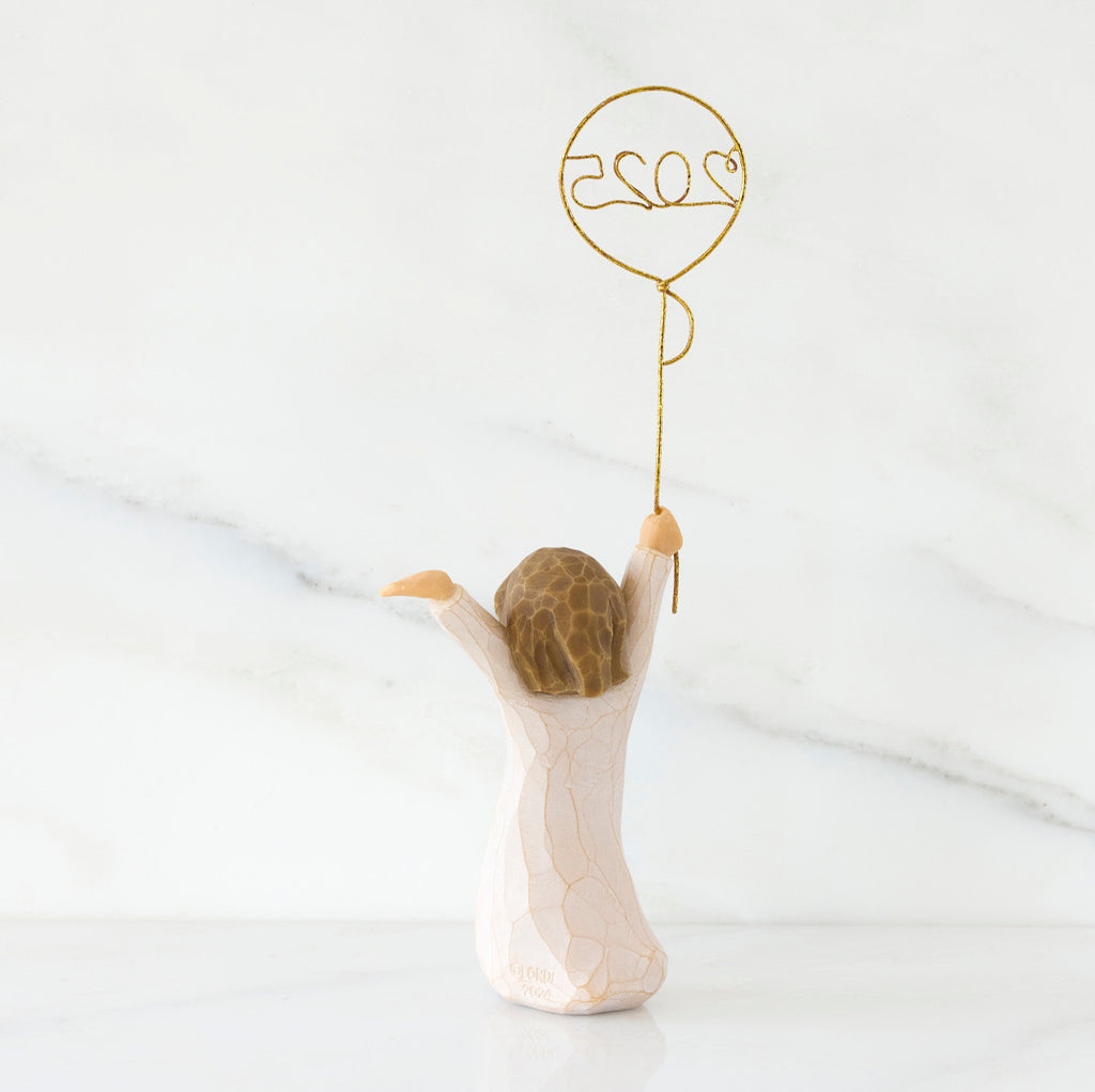 Back view: standing figure in cream dress, arms raised, holding gold wire balloon with 2025 written in gold wire inside