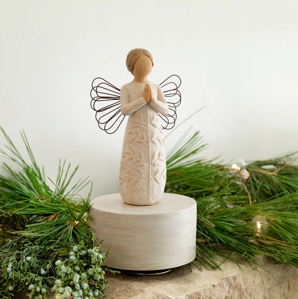 Willow Tree a tree, a prayer Musical, 7.5”h hand-painted resin angel figure in cream dress with wire wings, hands in prayer position, tree carving on dress, atop round, cream base containing musical element, on table with fir branches