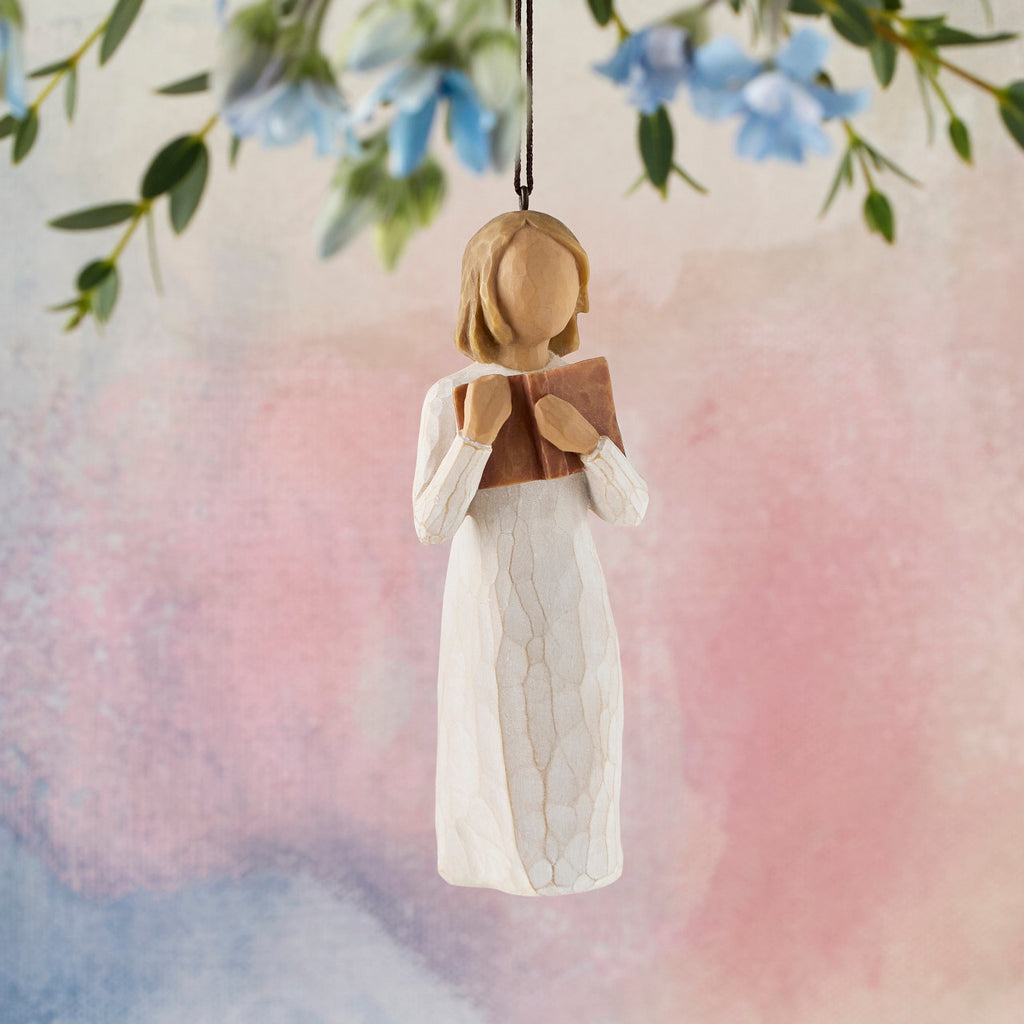 Willow Tree Love of Learning Ornament, 4”h hand-painted resin figure in cream dress holding open book to her chest, brown elastic cord, hanging in front of pink painting