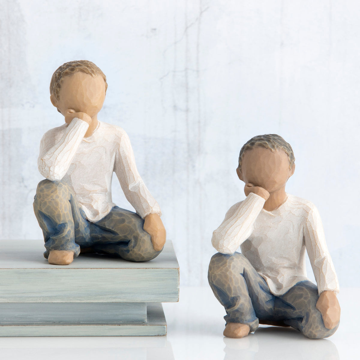 Two young brunette boy figurines in white and jeans sitting down with one arm to their face next to stack of books