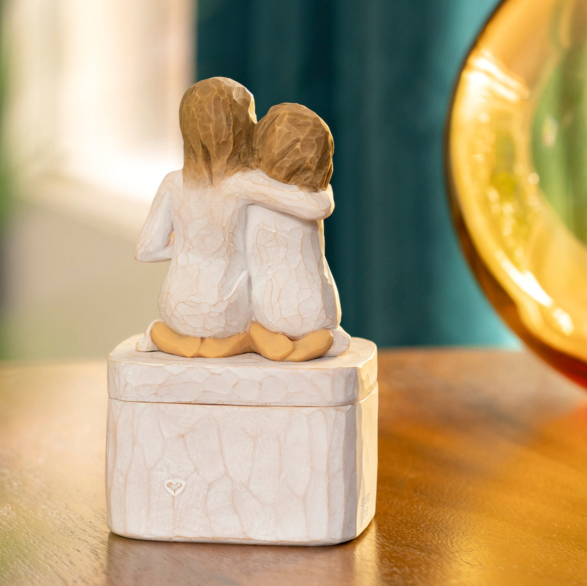 Willow Tree Sister mine Keepsake Box, back view of 3"sq hand-painted resin box with 3.5”H figure of two kneeling girls in cream dresses, arms around each other, affixed to lid, on wooden table